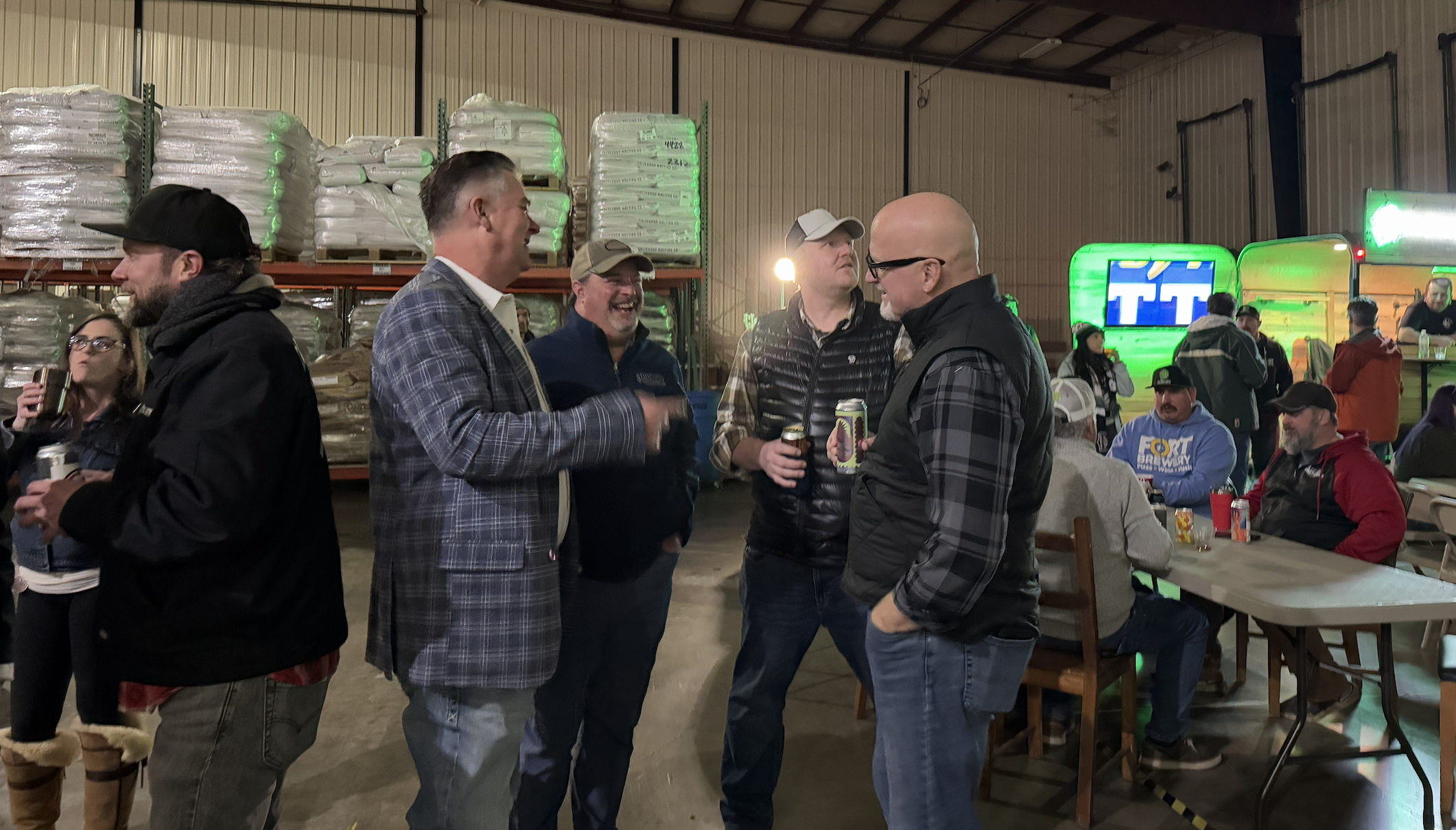 Group of people socializing at an indoor event, some holding drinks, in a warehouse-like setting with stacked supplies and a food truck in the background.