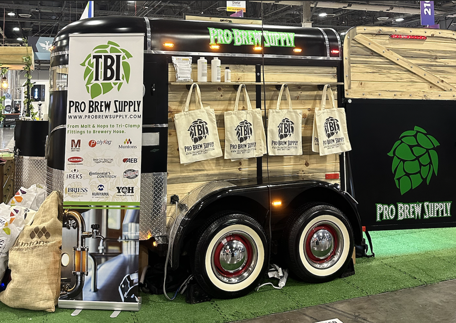 A mobile brewing supply trailer with wooden paneling, branding for Pro Brew Supply, and tote bags hanging on the side. There is a banner displaying product brands and a large hop logo, with artificial grass on the ground.