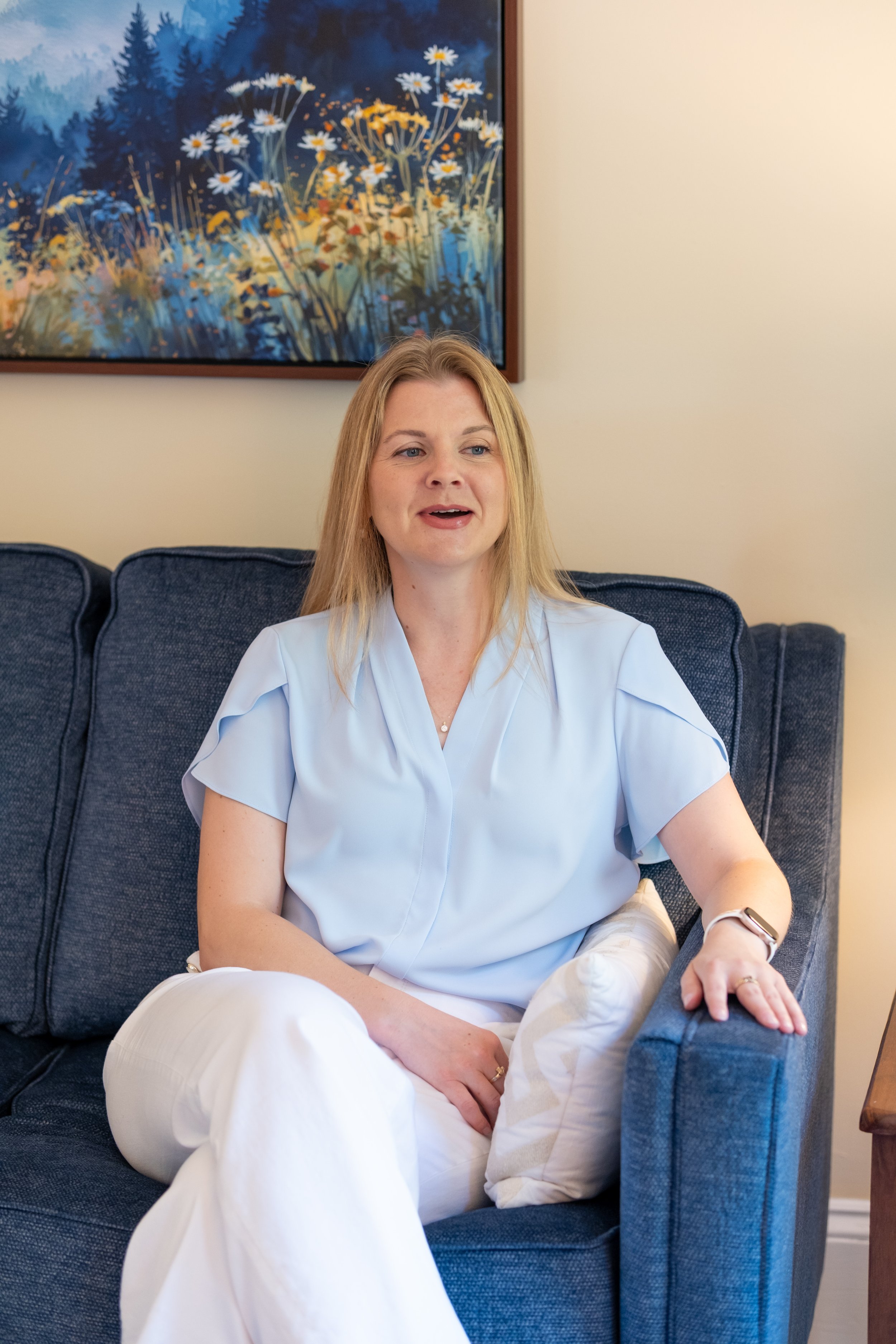 A woman with blonde hair sitting on a dark blue sofa, wearing a light blue blouse and white pants, with a colorful abstract landscape painting of flowers and mountains hanging on the wall behind her.