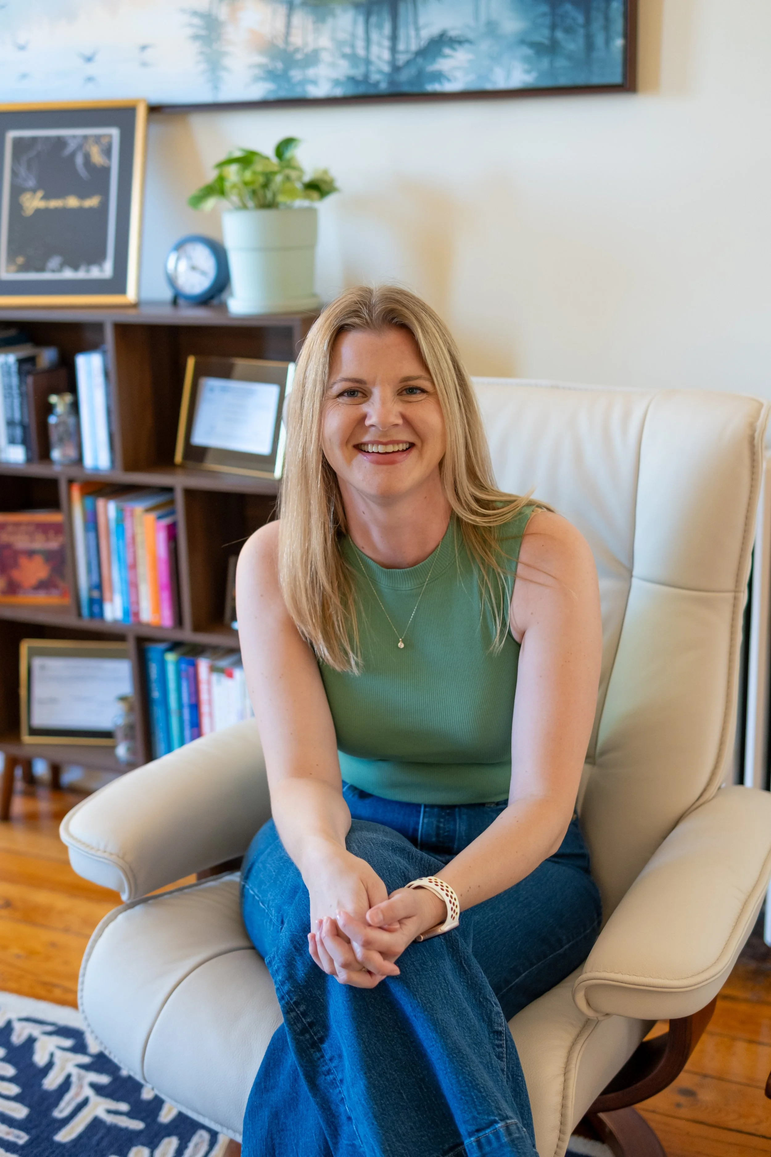 A woman with long blonde hair smiling, sitting in a beige armchair in a cozy living room with wooden floors, a bookshelf with books, a potted plant, and framed artwork on the wall behind her.