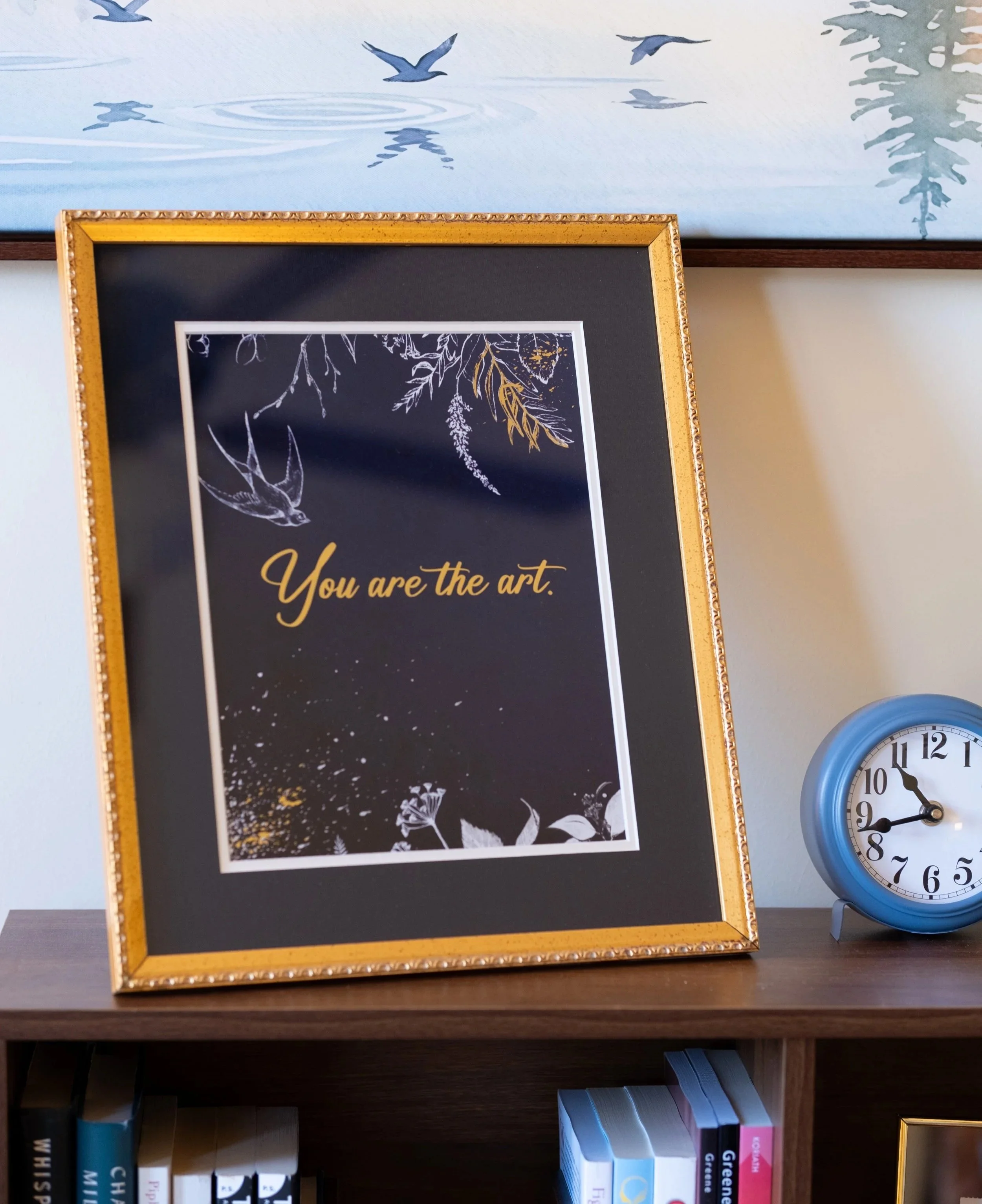 Shelf with books on the bottom, a framed quote on top that says 'You are the art,' and a decorative framed painting in the background, with a blue wall clock to the right.