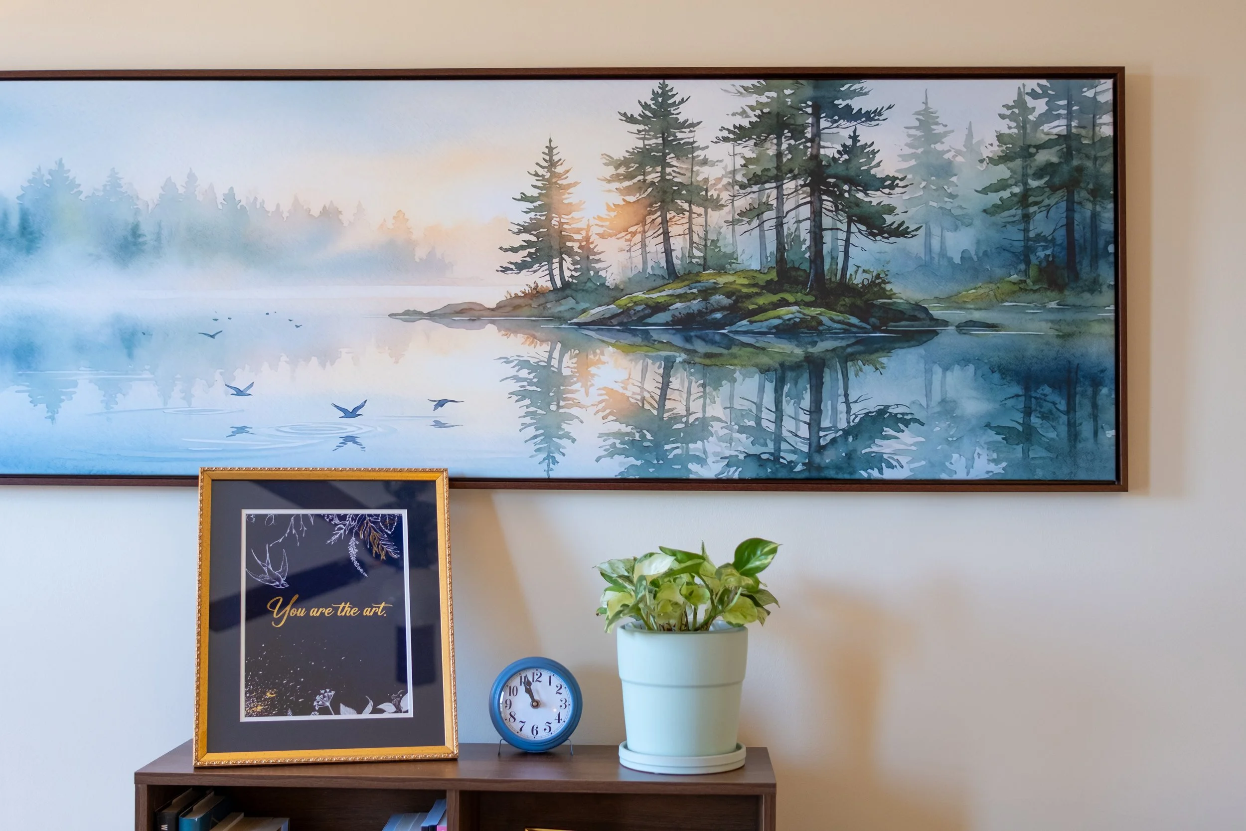 A framed artwork of a lake scene and trees leaning over water, hanging against a white wall above a bookshelf with framed art, a clock, and a potted plant on top.