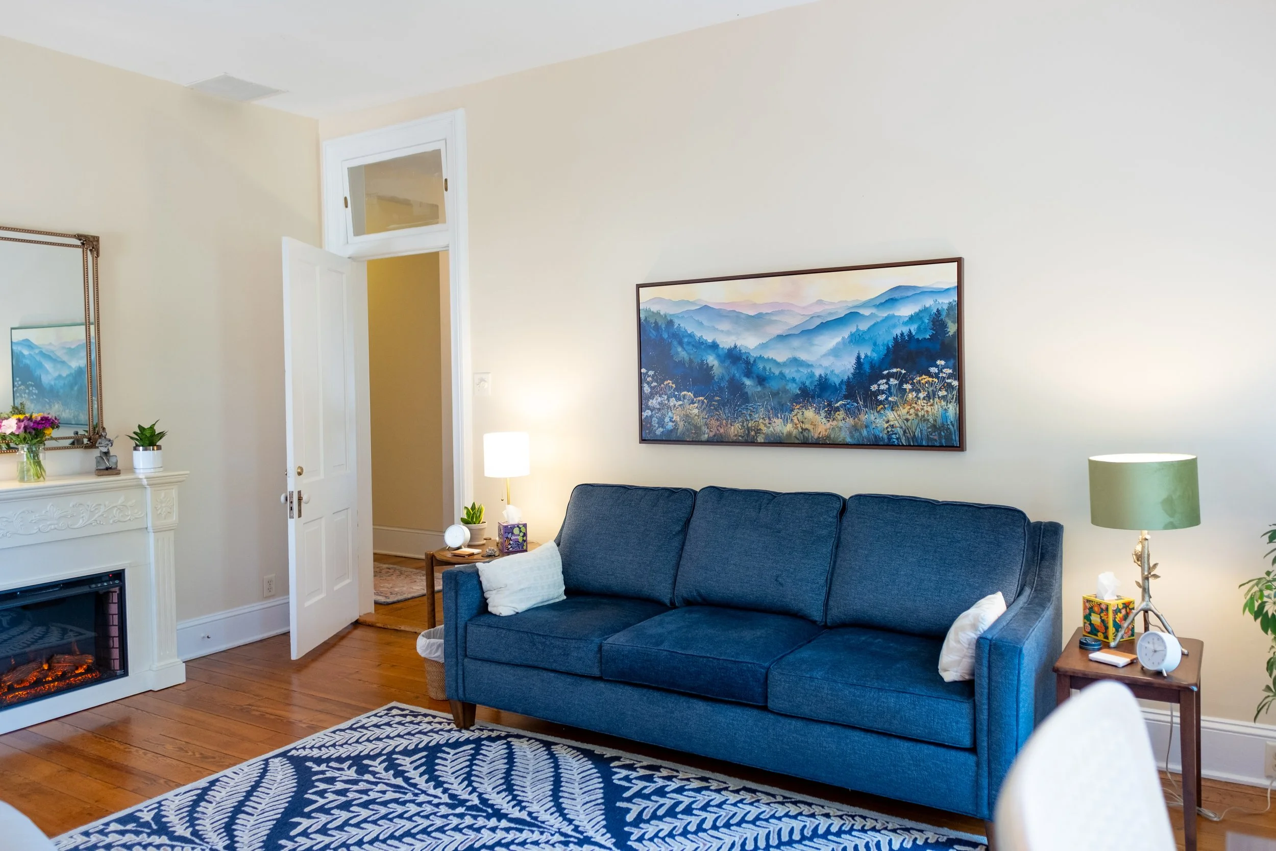 Comfortable counseling room with a blue sofa, a wooden side table with a clock, framed landscape painting on cream wall, fireplace with decorations, and wooden floor.