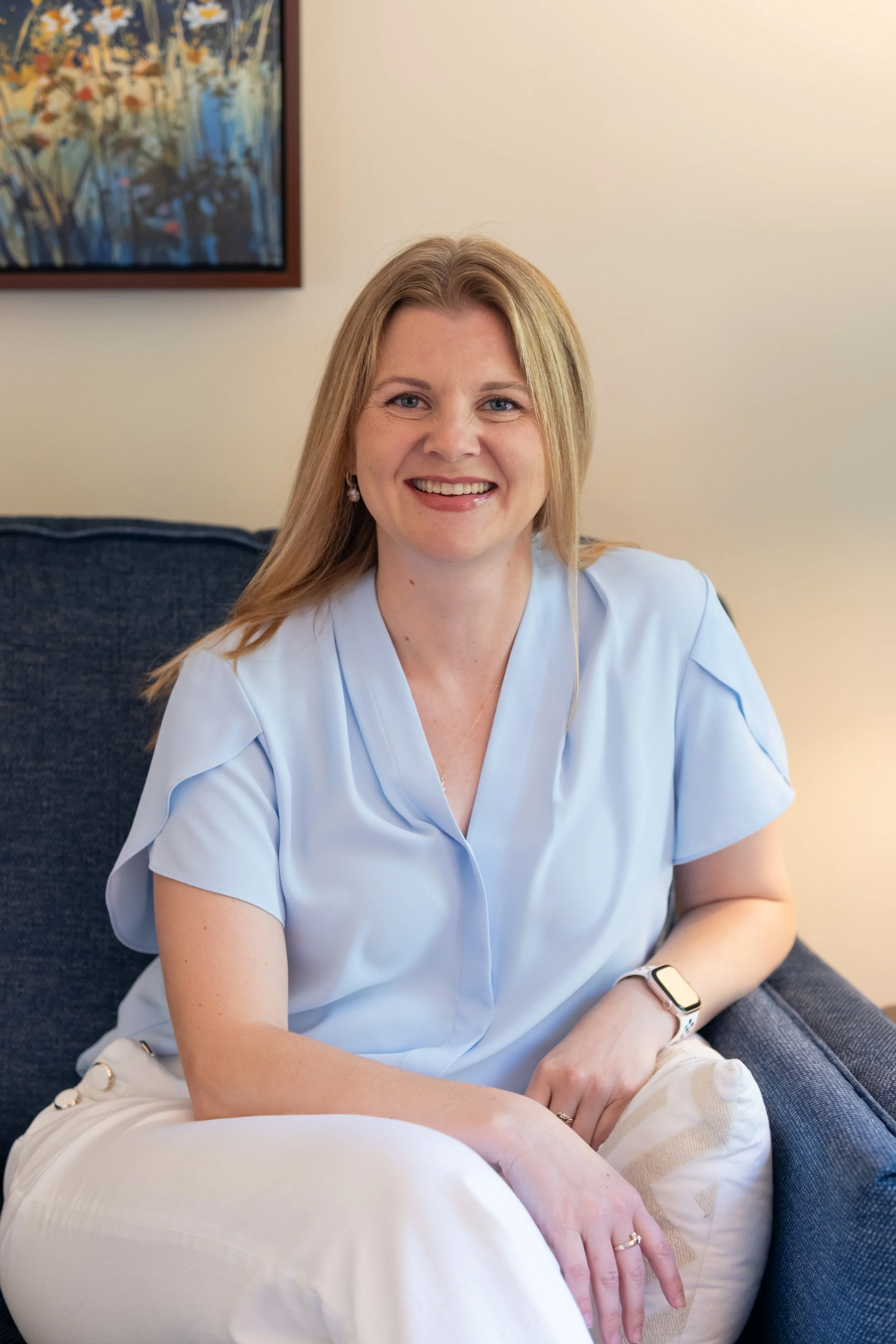 A woman with long blonde hair, wearing a light blue shirt and earrings, smiling while sitting on a dark-colored couch with her arm resting on her lap. There is a colorful painting on the wall behind her.