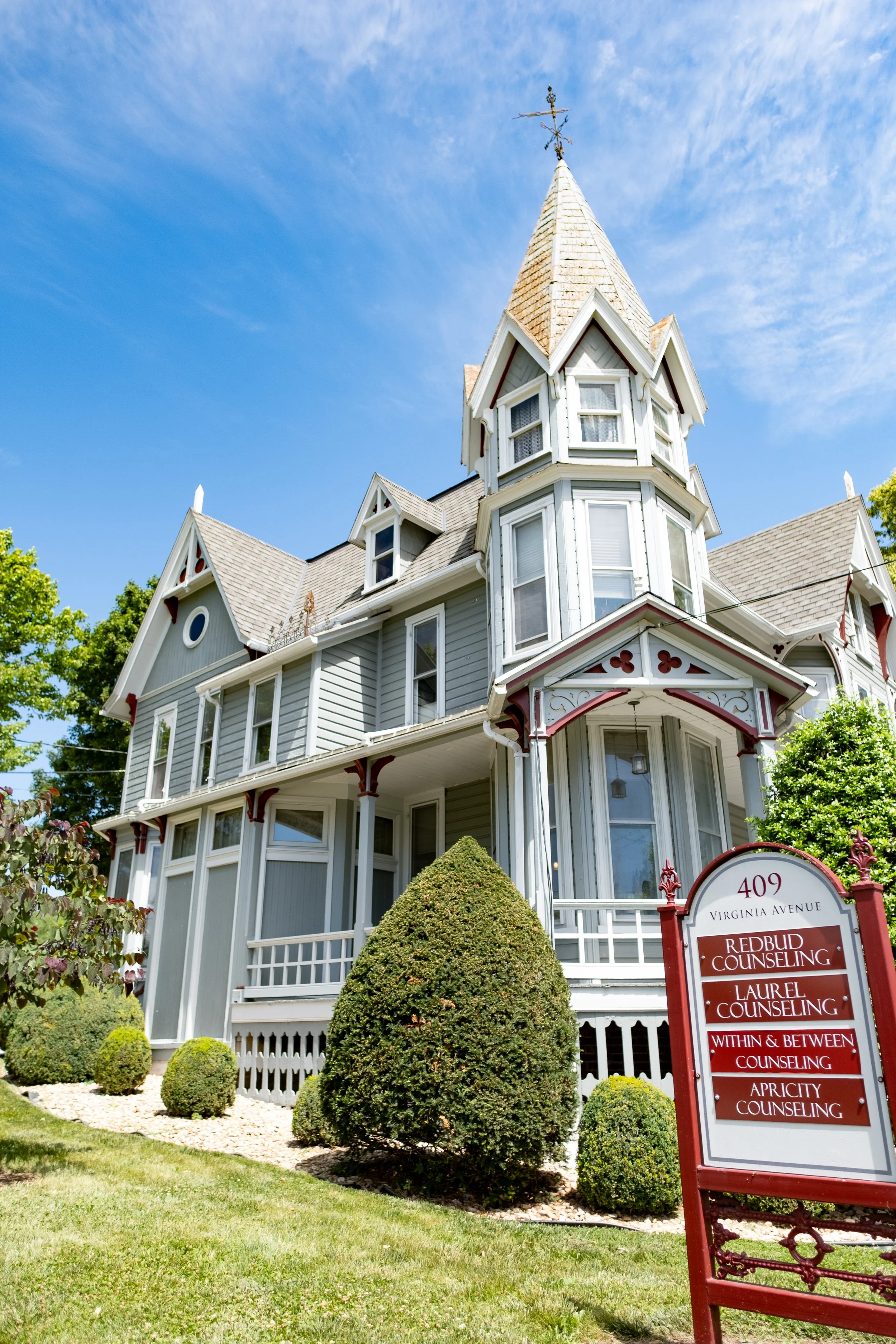 A Victorian-style house with a prominent tower and turret under a blue sky with scattered clouds, surrounded by a well-kept lawn and bushes. A sign in front of the house lists counseling services.