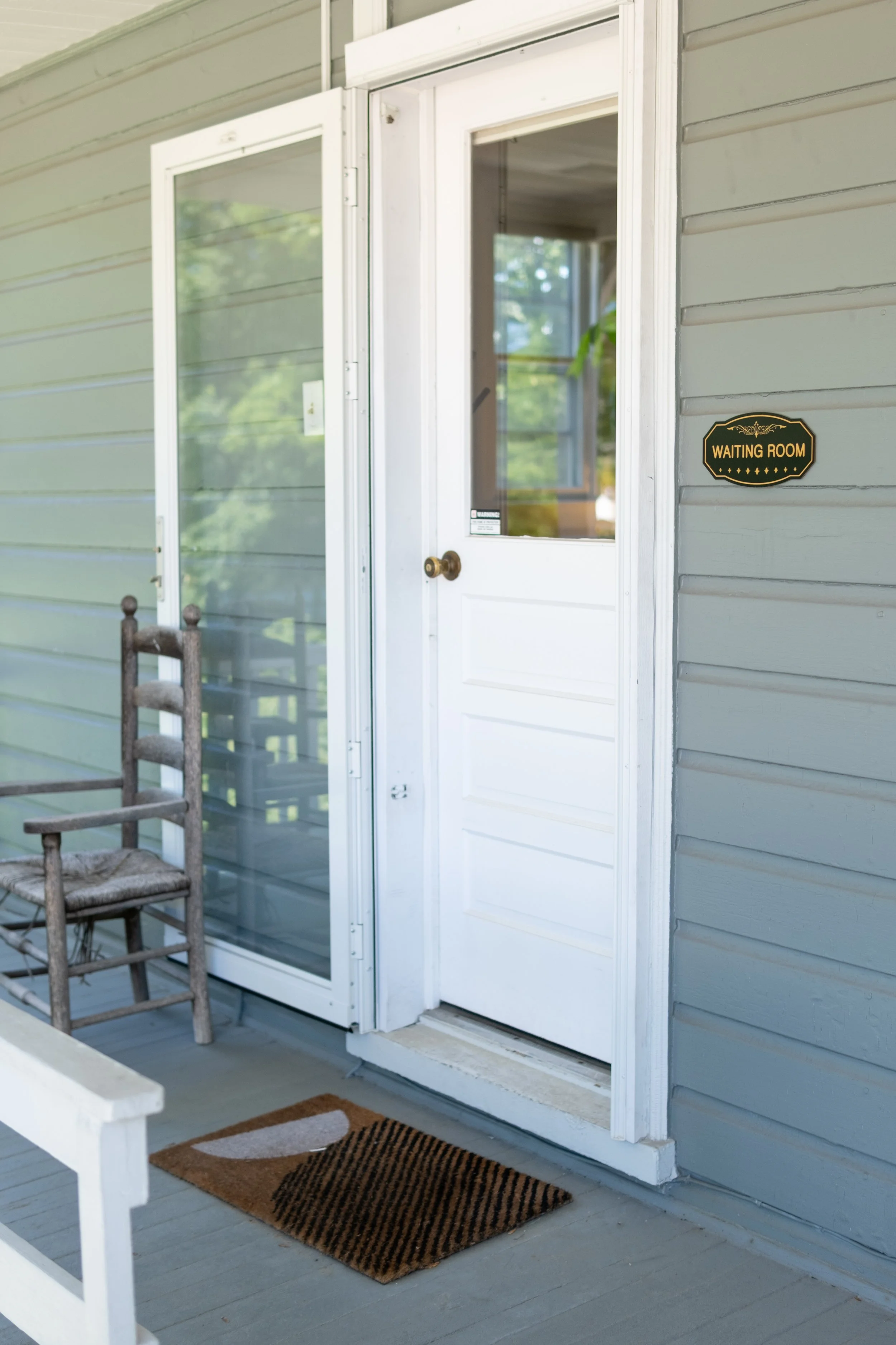 Exterior porch with a white screen door, grey siding, a wooden chair, a welcome mat, and a black sign that reads 'Waiting Room'