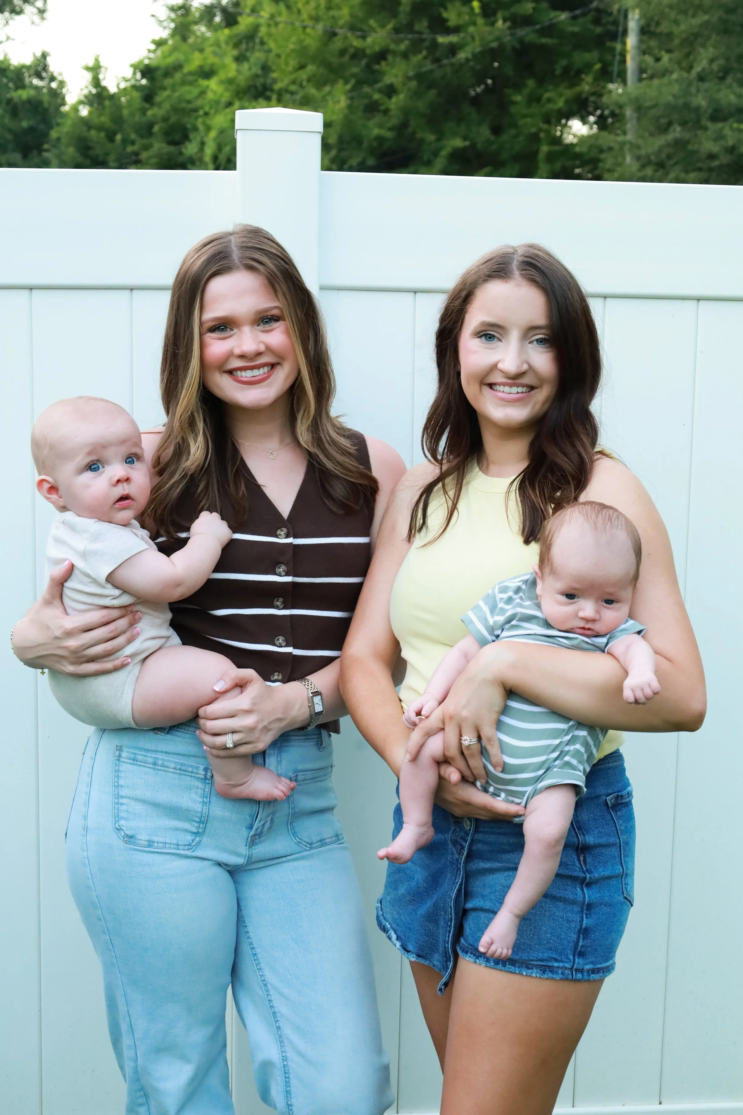 Two women holding babies in front of a white fence with green trees in the background.