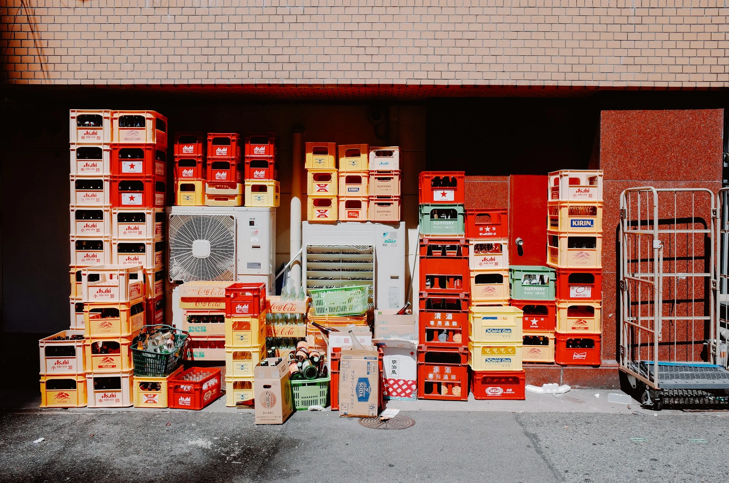 Stacked plastic crates and boxes filled with bottles, cans, and miscellaneous items under an overhang against a brick wall.