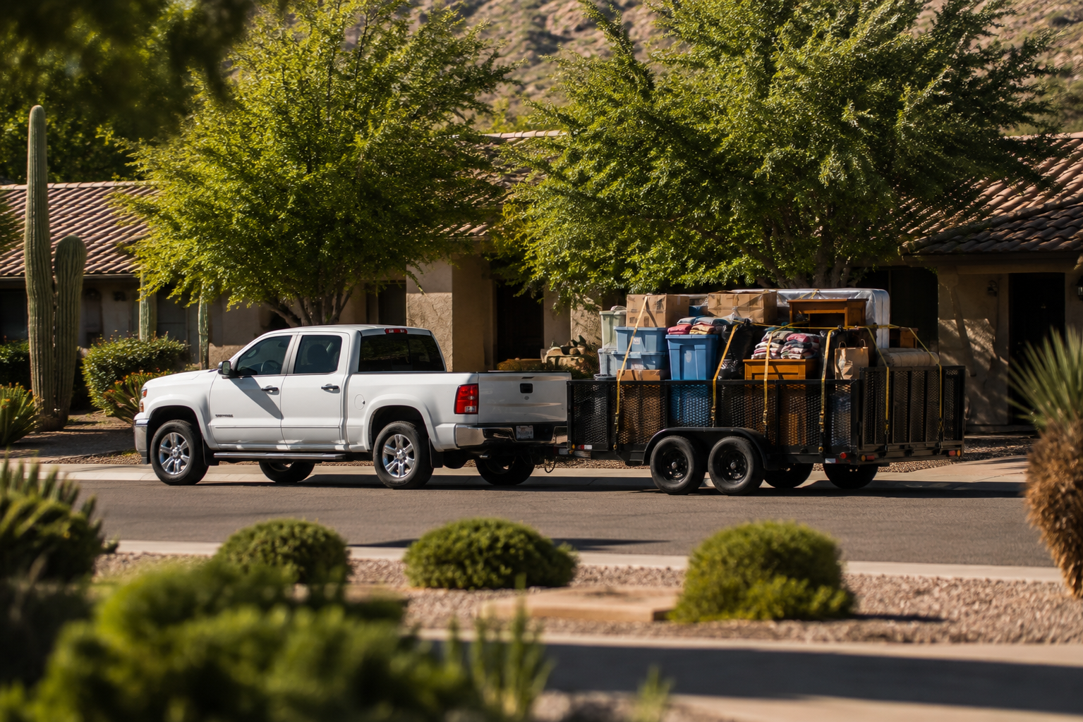 A white pickup truck pulling a trailer loaded with boxes, bags, and furniture down a suburban street with trees and bushes.