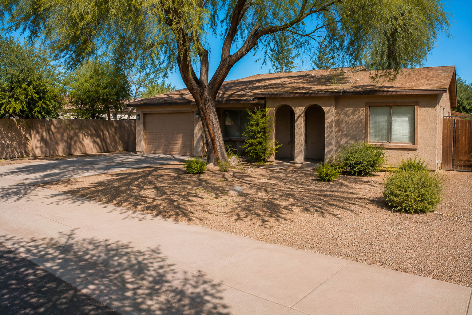 A single-story house with a light brown exterior, a brown tiled roof, and a two-car garage. In front of the house, there are desert landscaping with a large tree casting shadows, small shrubs, and rocks. The sidewalk runs along the front of the yard, and the sky is clear and blue.