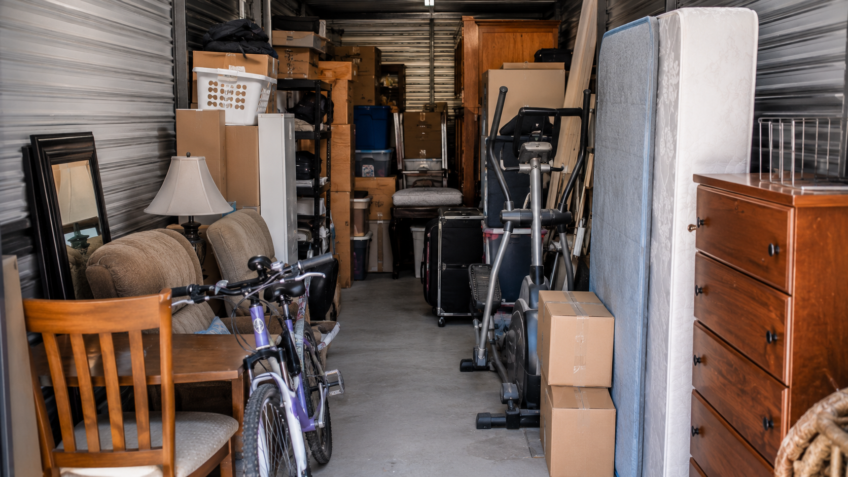 Storage unit filled with various household items including furniture, a bicycle, exercise equipment, boxes, a lamp, and mirrors.