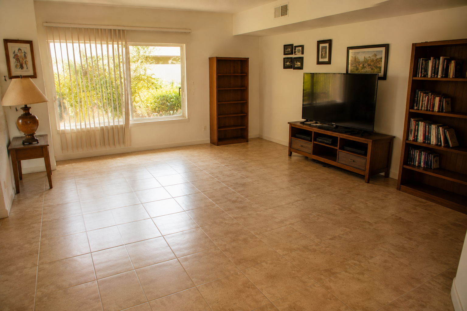 Empty living room with tiled floor, large window with vertical blinds, and wooden furniture including bookshelves and TV stand.