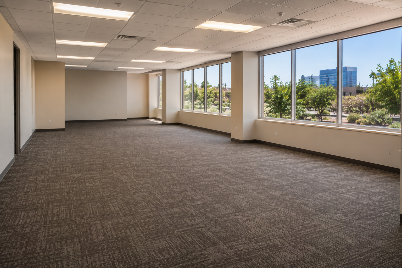 Empty office space with large windows, beige walls, and brown carpet, overlooking trees and buildings outside.