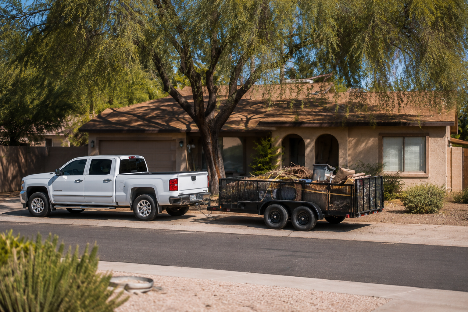 White pickup truck hitched to a trailer filled with yard debris parked in front of a beige house with a tree providing shade.