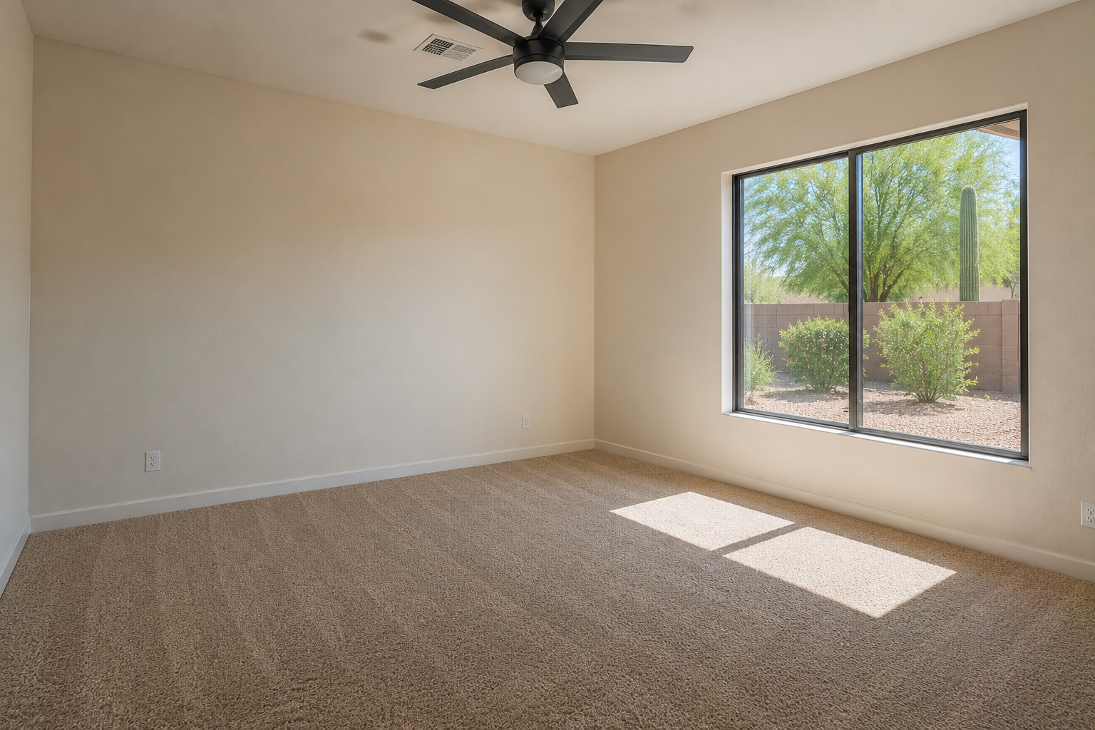 Empty room with beige carpet, beige walls, large window showing desert landscape with trees, bushes, cactus, ceiling fan