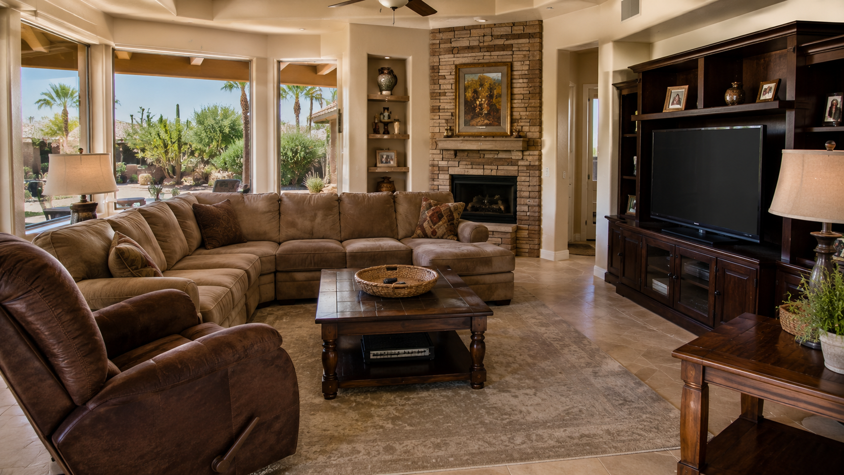 Living room with large beige sectional sofa, brown leather recliner, coffee table, and wooden entertainment center with television. Large windows show outdoor patio and desert landscape with palm trees.