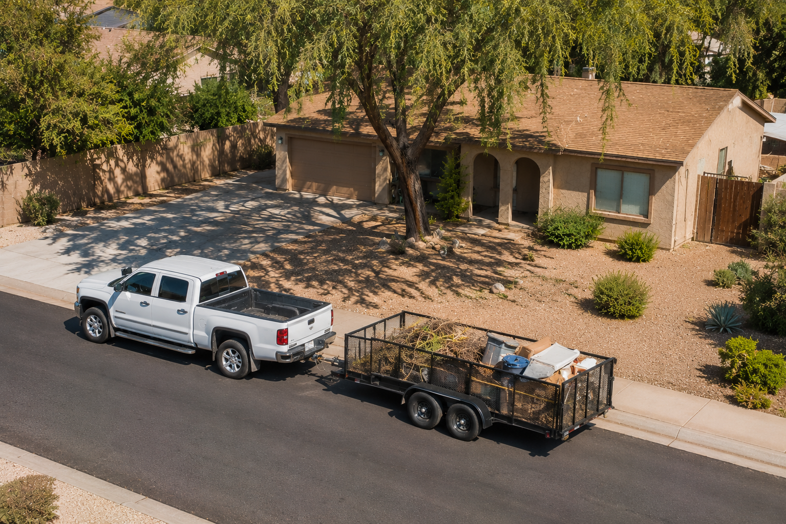 A white pickup truck is parked alongside a house with a brown roof, dragging a trailer filled with assorted items. The house has a front yard with desert landscaping, small bushes, and a large tree casting shadows on the yard and driveway.