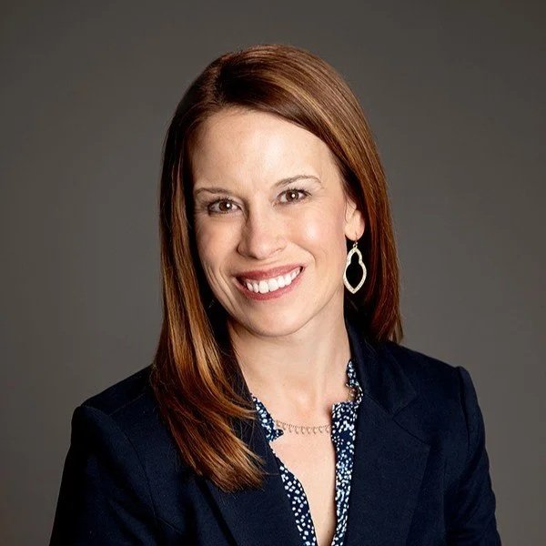 Megan Ryan wearing a navy blazer, patterned blouse, and large earrings, smiling against a gray background.