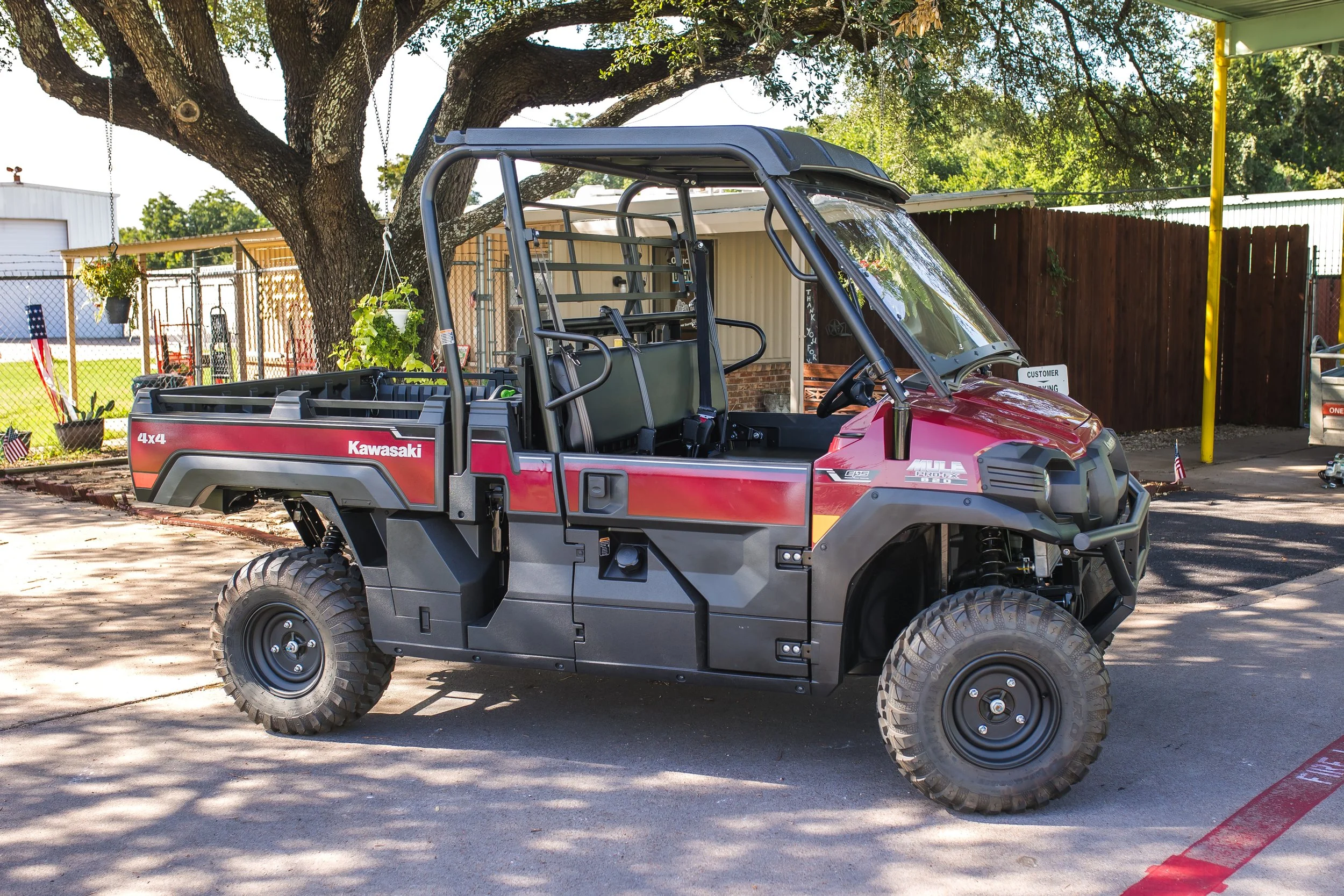 A red Kawasaki utility vehicle with black tires parked outdoors under a large tree with green foliage. The vehicle has an open cargo bed at the back and a canopy roof. There is a fenced area and a building in the background.