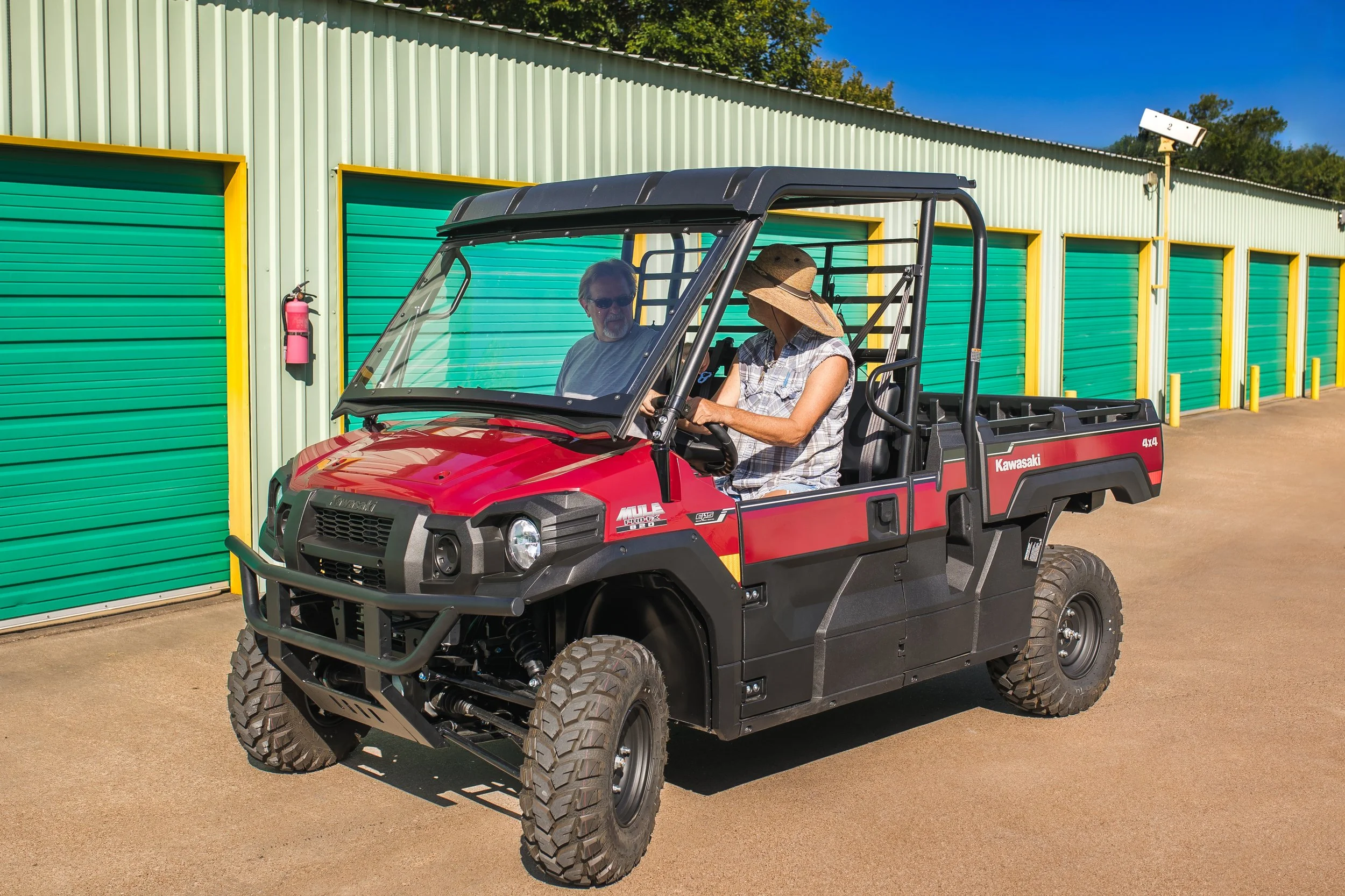 A man and a woman riding a red Kawasaki utility vehicle outside a storage facility with green garage doors and a fire extinguisher mounted on the wall.