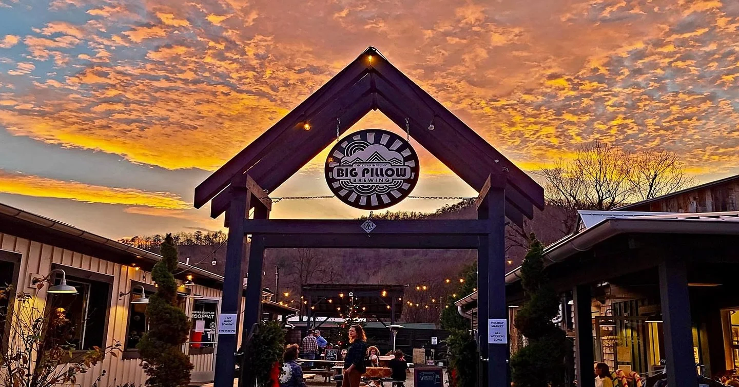 Outdoor scene at sunset with a dark blue building and string lights, people sitting at tables, and a large round sign reading 'Big Pillow Brewing' hanging under a gabled roof, with a colorful sky in the background.