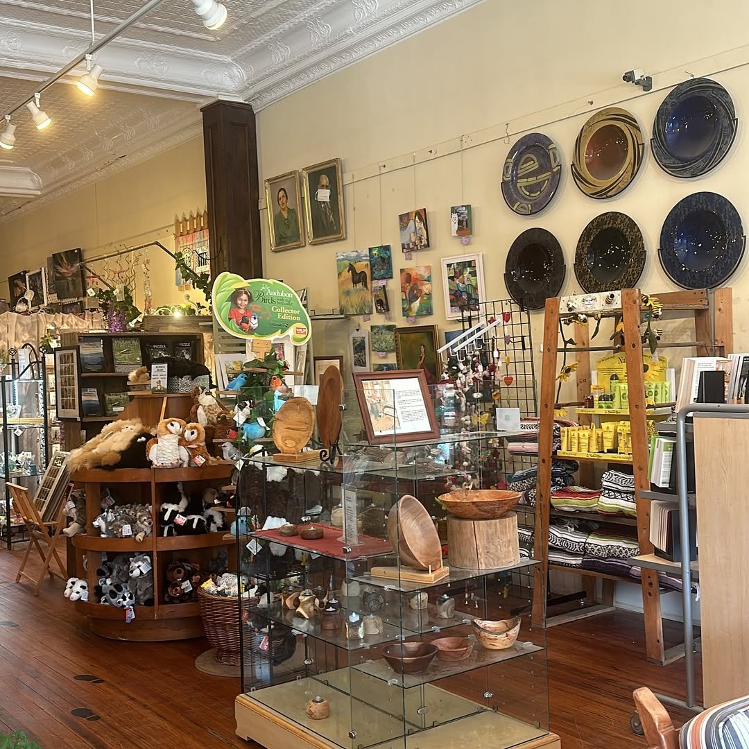 Interior of an antique or gift shop with shelves and display cases filled with plush toys, pottery, artwork, and decorative items. The walls are decorated with framed paintings and circular metal trays, with track lighting on the ceiling.
