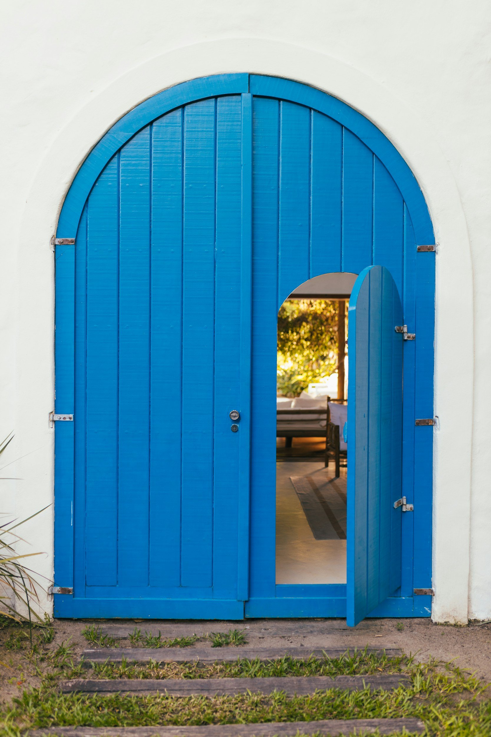 Open blue wooden arched door with a small window, showing a patio with furniture and trees outside.