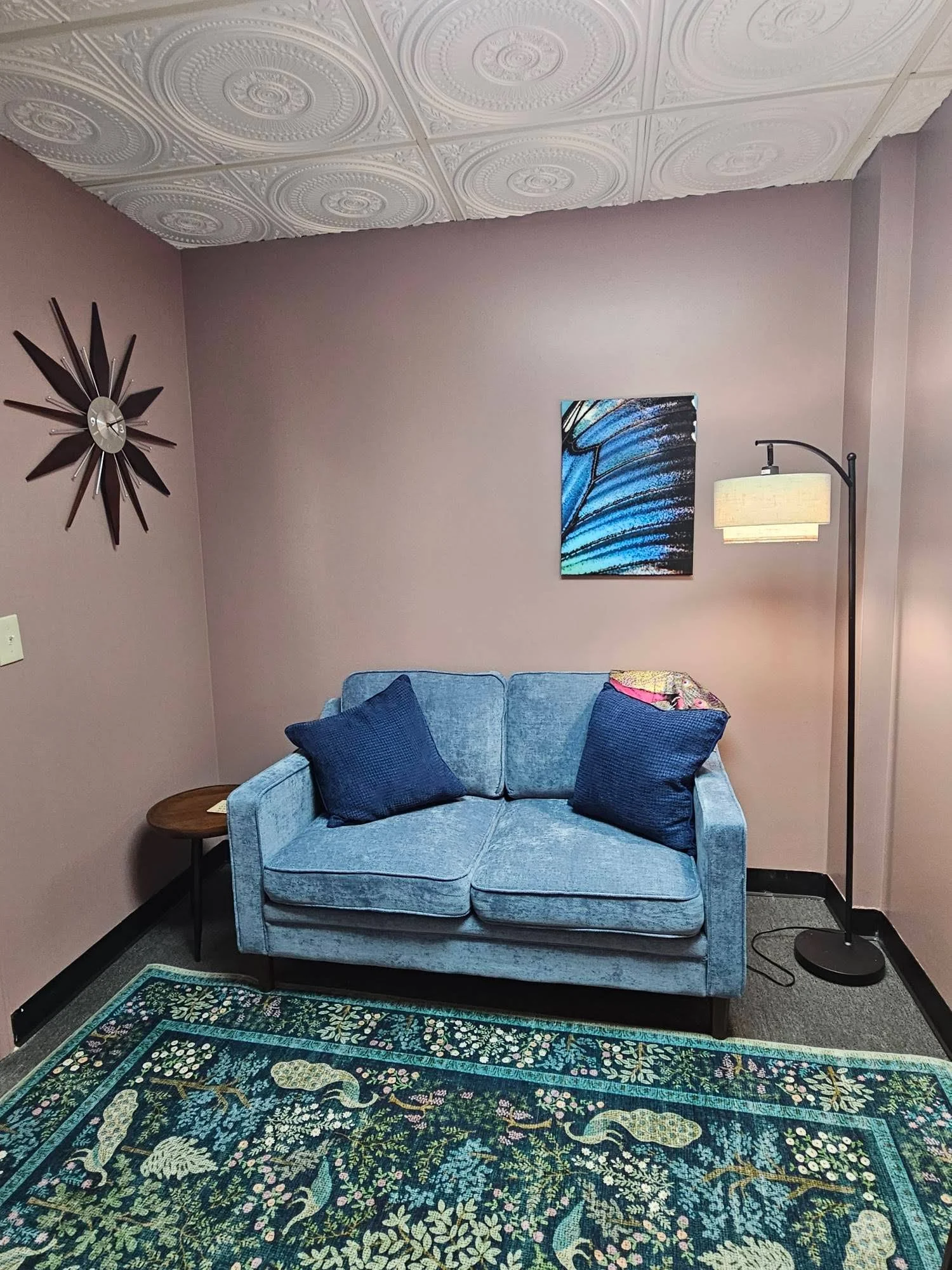 A cozy living room corner with a blue upholstered loveseat, navy blue pillows, a wooden side table, a colorful abstract painting on the wall, a tall floor lamp with a cream lamp shade, a decorative area rug with a floral and animal motif, and a unique starburst clock on the mauve wall.