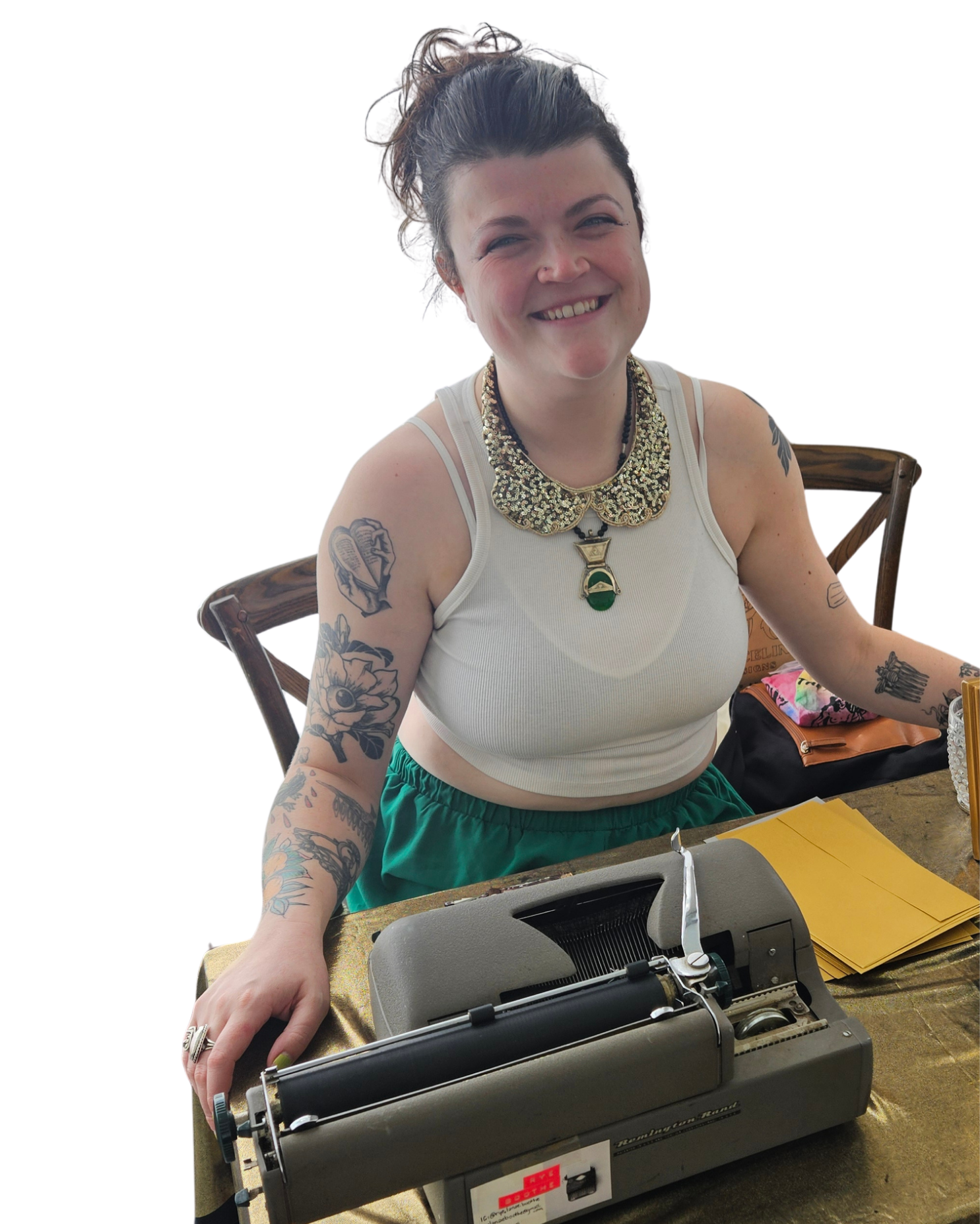 A smiling woman with tattoos on her arms, wearing a sleeveless white top, sits at a table with yellow folders and a vintage typewriter in front of her. She has short, wavy hair and a chunky necklace with a pendant. The background appears to be a home or office setting.