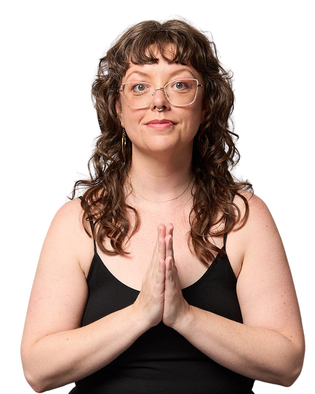 A woman with curly brown hair, glasses, and a septum piercing, wearing a black tank top, holding her hands together in prayer or greeting pose, looking content.