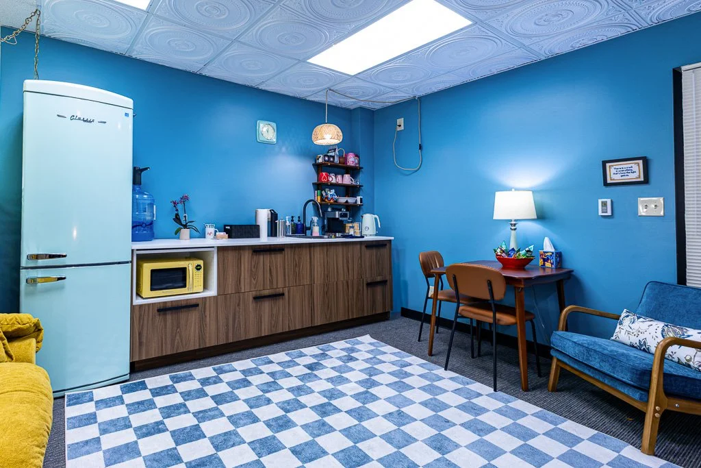 Small kitchen area with a vintage refrigerator, water dispenser, microwave, and shelving; adjacent to a dining area with a table, chairs, lamp, and a blue sofa.