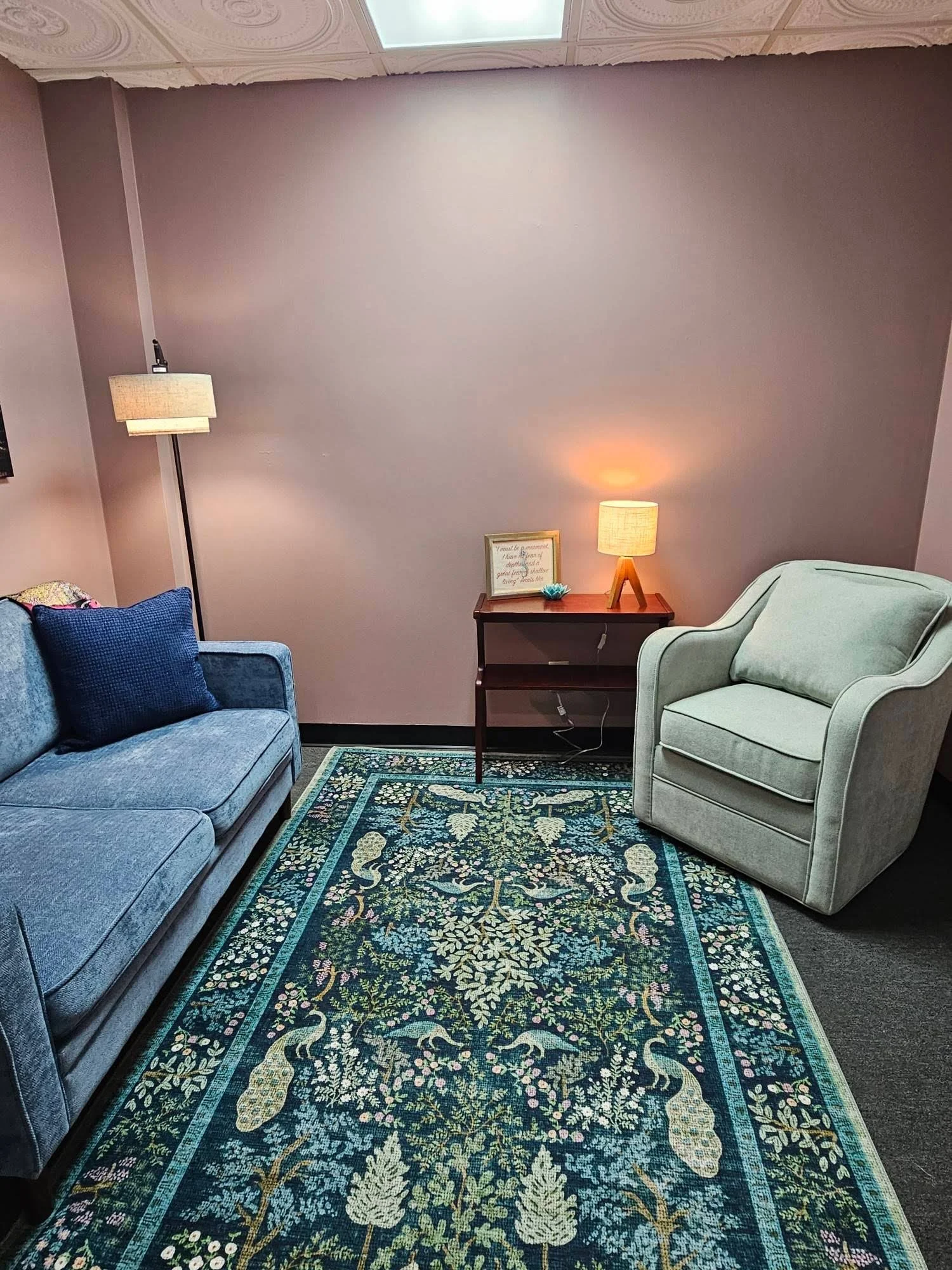 A living room with a floral rug, a blue upholstered sofa with navy pillows on the left, an armchair on the right, and a small side table with a lamp and framed artwork, illuminated by warm lighting and surrounded by pinkish walls.