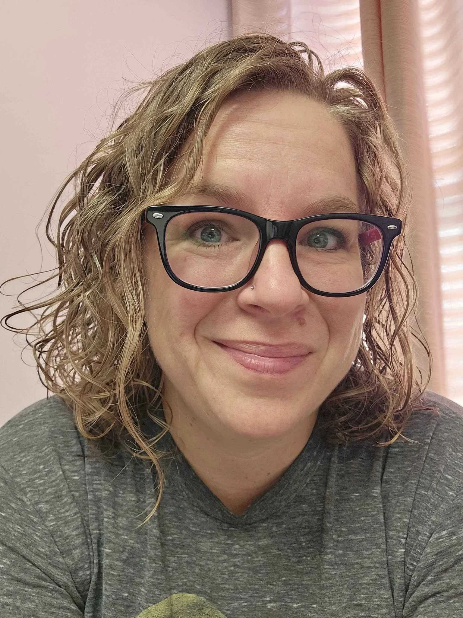 Close-up of a woman with curly blonde hair, glasses, and a nose piercing, smiling in front of pink curtains.