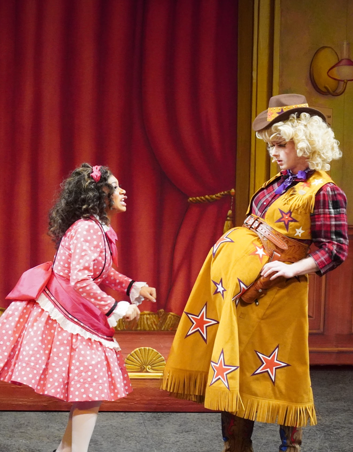 A scene from a play featuring two women dressed in colorful costumes, with red curtains and a wooden stage in the background. One woman is wearing a pink polka-dot dress with a matching bow, and the other is wearing a cowboy-themed outfit with a star