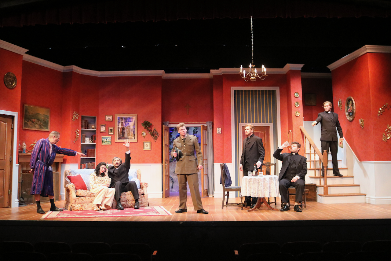 Actors perform a scene on stage resembling a cozy living room with red walls, framed pictures, and a staircase. Characters are dressed in period costumes, with some seated on a floral sofa and others standing or sitting at a table, engaged in acting.