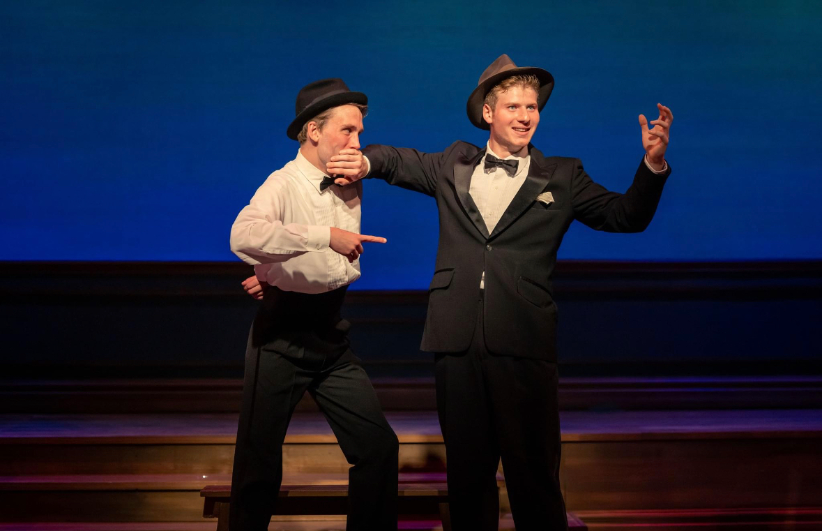 Two men dressed in tuxedos, hats, and bow ties stand on a stage with a blue background. One man muffles the other man's mouth while the second man gestures with his hand, suggesting a theatrical performance.