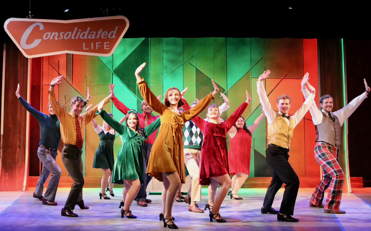 The image shows a group of actors performing a lively musical on stage, dressed in vintage costumes, with a colorful geometric background.