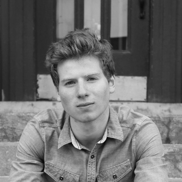 A young man with wavy hair sitting on steps outside, wearing a button-up shirt, looking directly at the camera.
