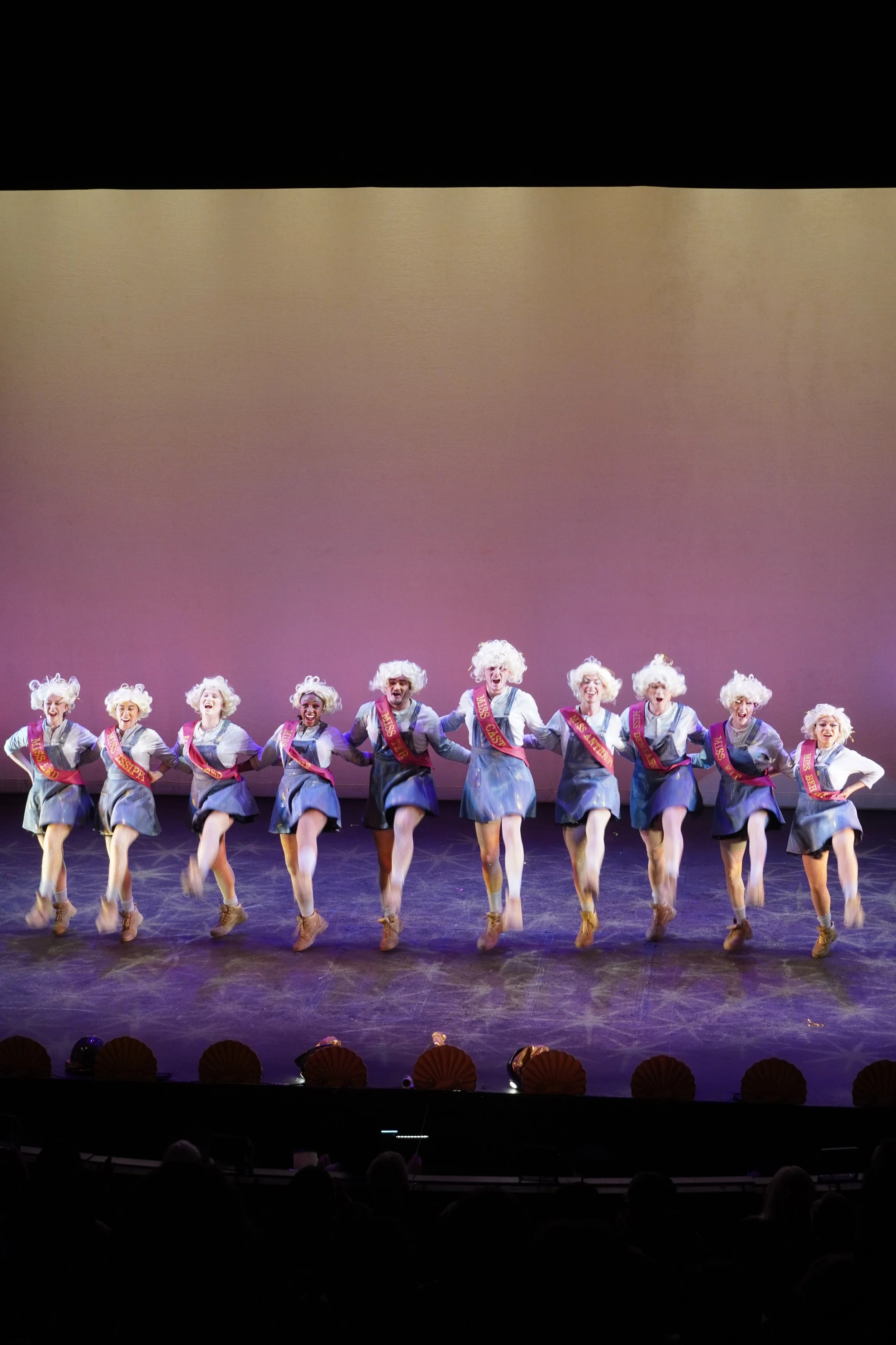 Group of women dressed in costumes with sashes performing a tap dance on stage, holding hands, with an audience watching.