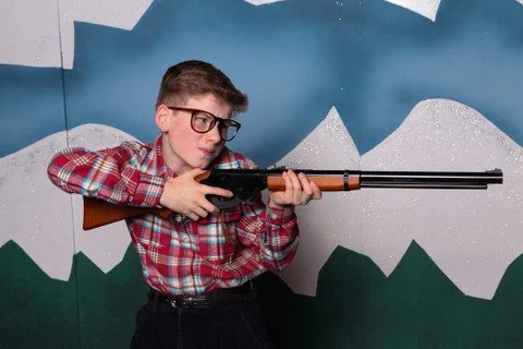 A young person wearing glasses and a plaid shirt aiming a rifle in front of a decorated background.