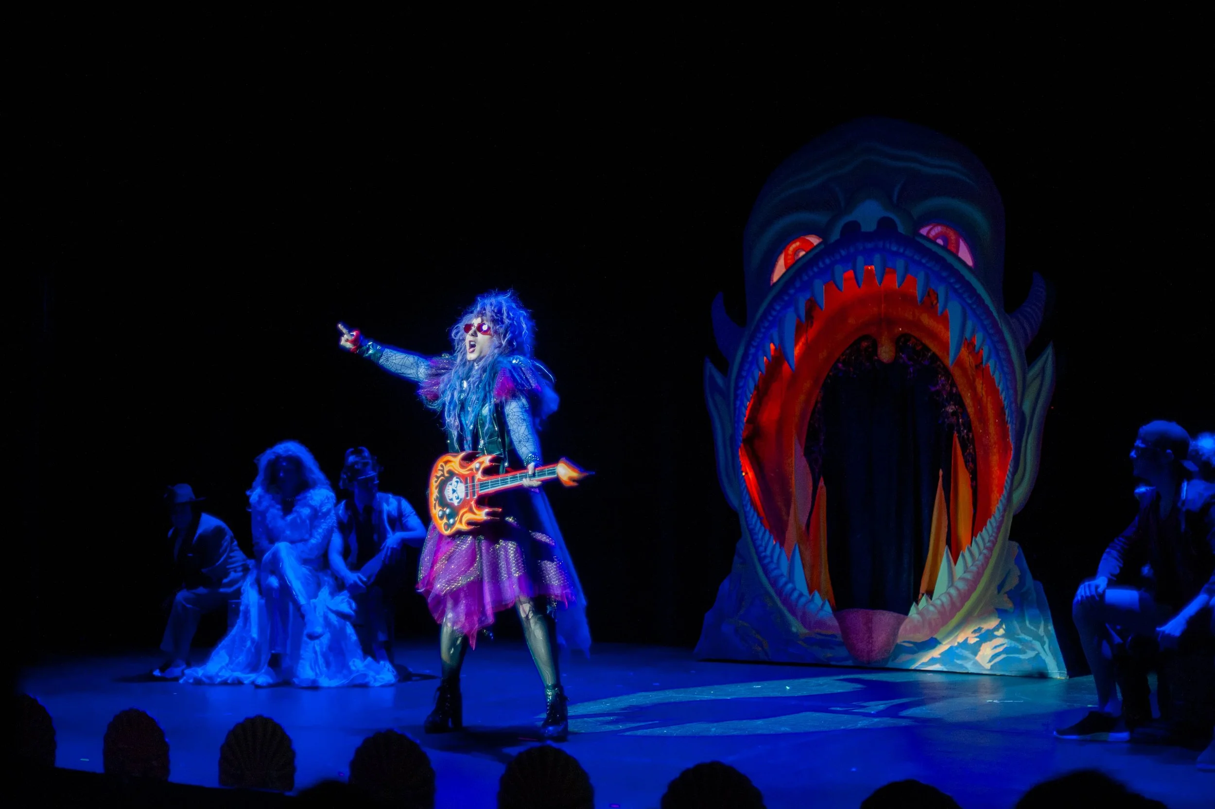 Performer in colorful costume holding a guitar, singing on stage with a large brightly lit dragon puppet in the background, surrounded by other performers in costumes.