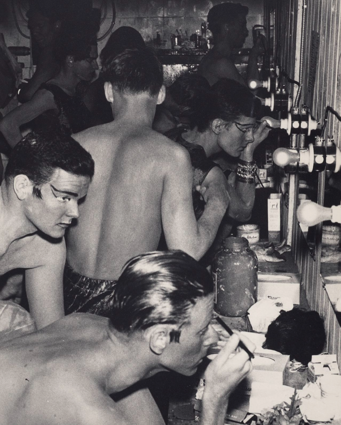 A black and white photo of several young people, mostly shirtless, working at a laboratory or workshop bench with scientific equipment, bottles, and supplies.