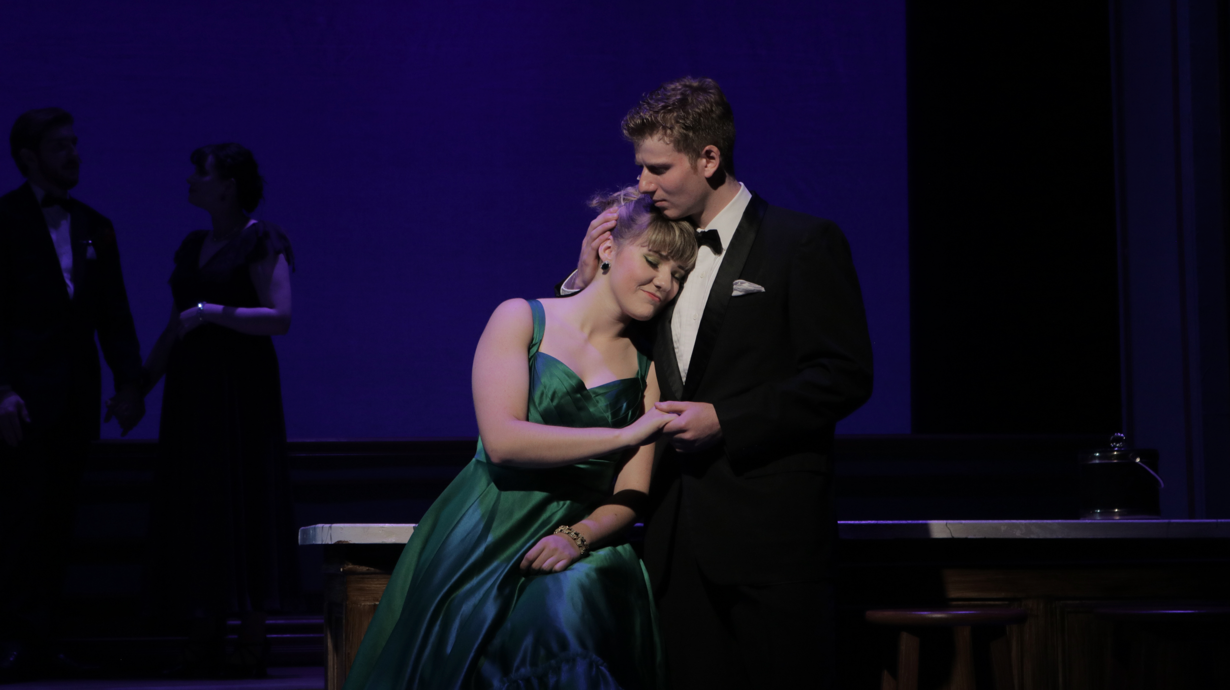 A young man and woman dressed in formal attire embrace on stage during a theatrical performance; the woman is wearing a green dress, and the man is in a black tuxedo. In the background, two other performers in elegant dress are holding hands.