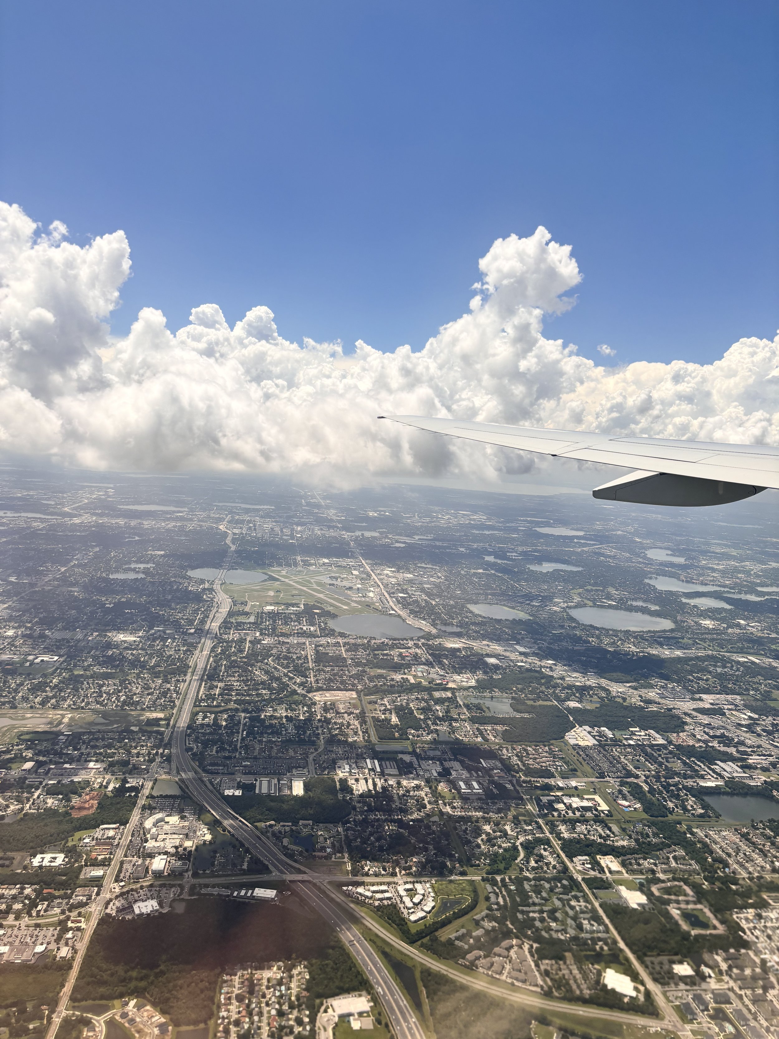 Aerial view of a city with roads, lakes, and buildings seen from an airplane window, showing blue sky with large clouds.