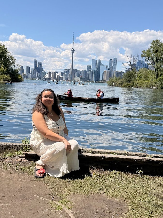 A woman sitting by a river with a city skyline, including the CN Tower, in the background; there are boats on the water and greenery on the sides.