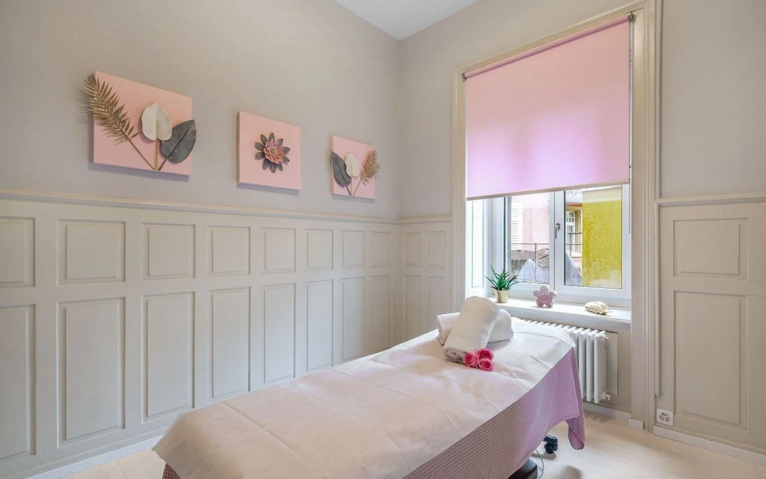 Schlafzimmer mit Bett, Wände mit Holzpaneelen, Fenster mit pinkem Rollos, Wanddekoration mit Blumenmotiven in Pastellfarben