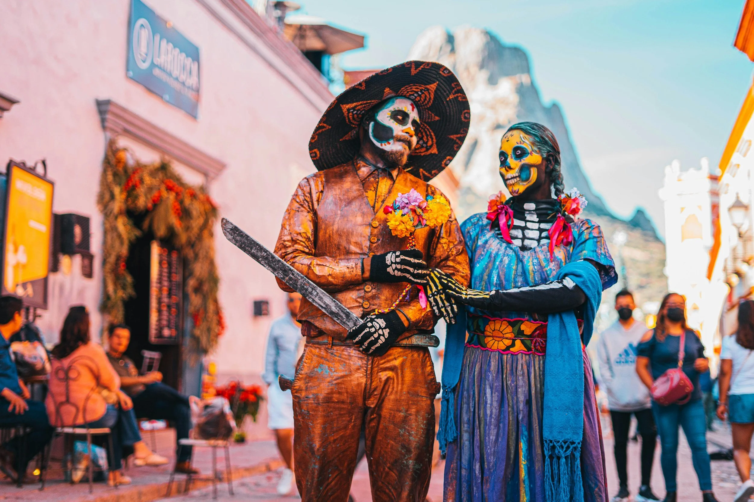 Two people dressed as skeletons and wearing traditional Mexican Day of the Dead face paint, with one person wearing a large black hat, embrace each other on a busy street during a celebration or festival.