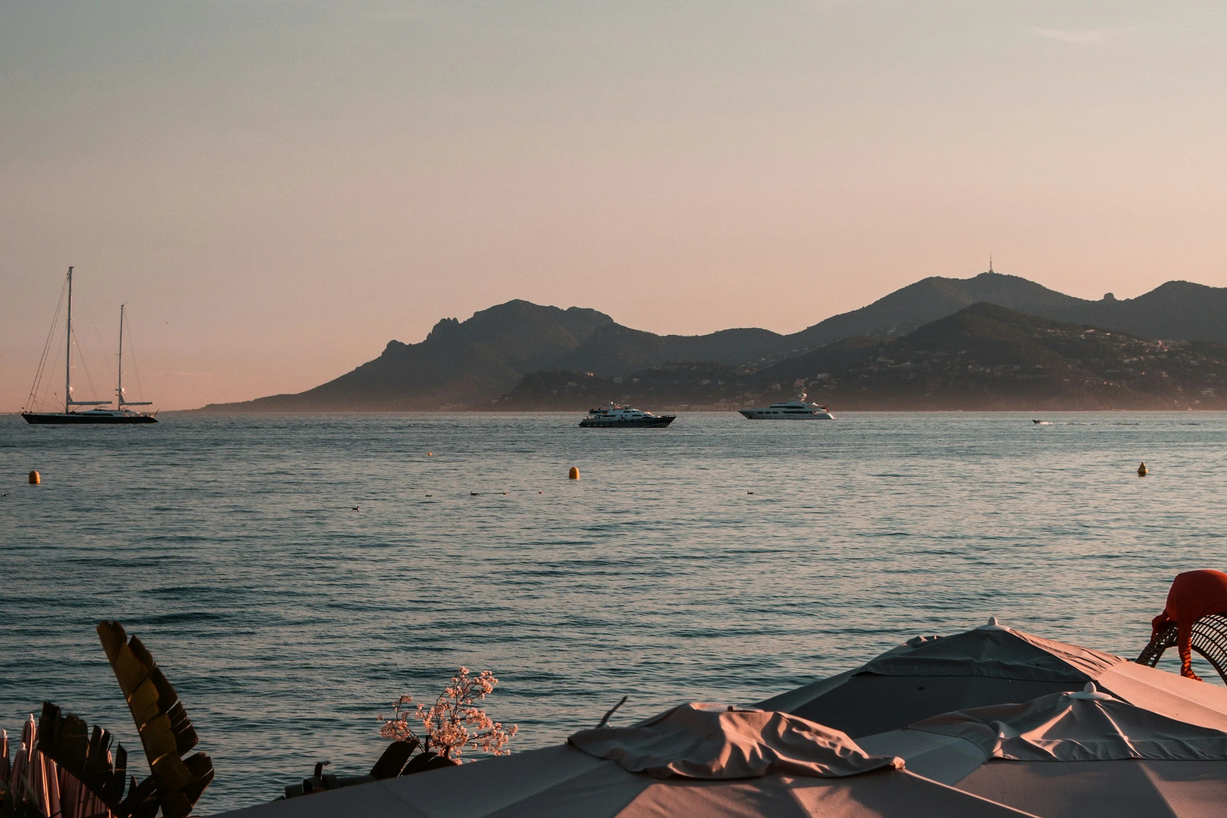 A scenic view of a calm ocean with three boats floating on the water and mountainous terrain in the background, during sunset or sunrise.
