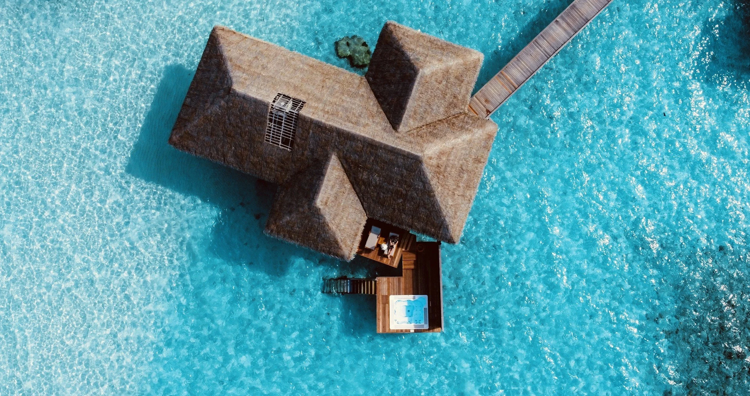 Aerial view of a thatched-roof overwater bungalow hotel with a private deck, jacuzzi, and a walkway extending into clear blue water.