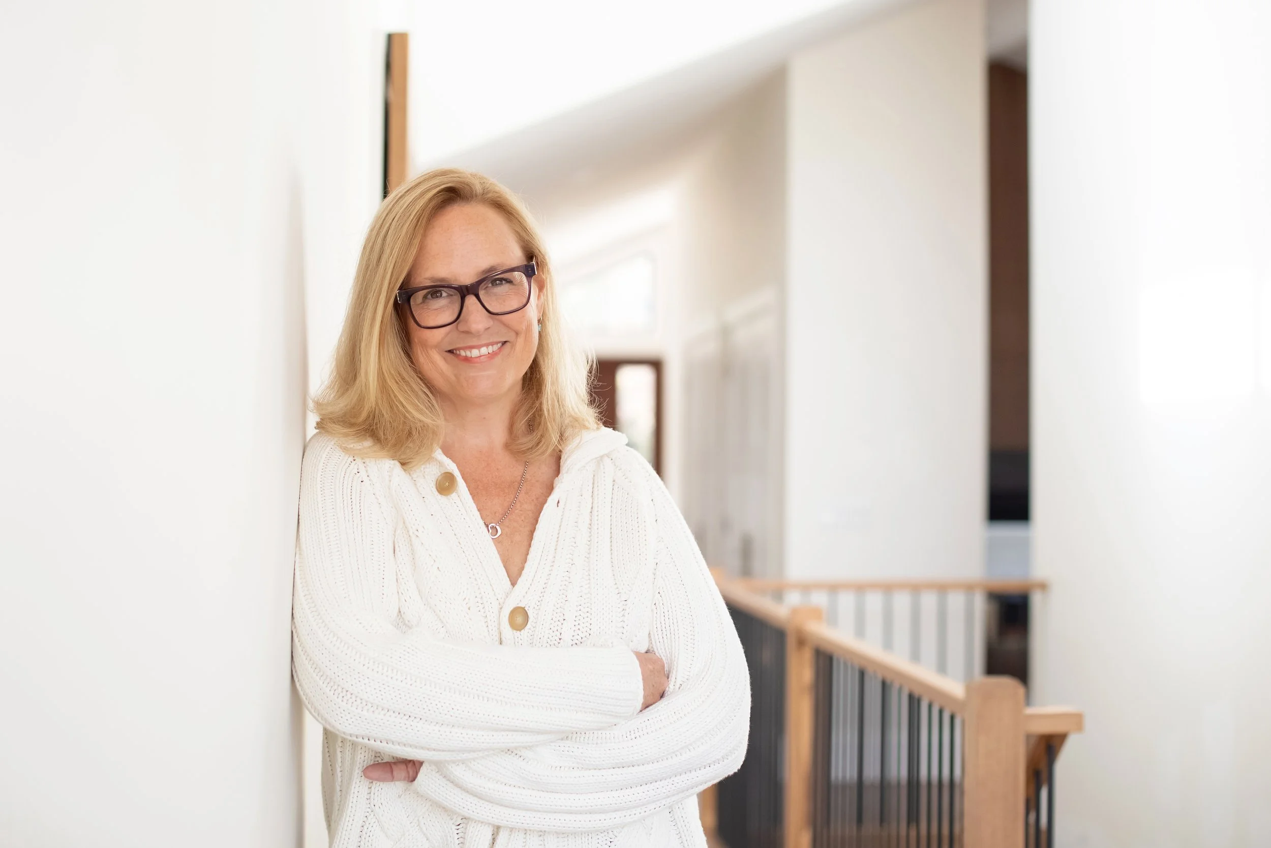 A woman with blonde hair, glasses, and a white cardigan smiling and standing with arms crossed in a bright, modern interior space with a wooden staircase railing.